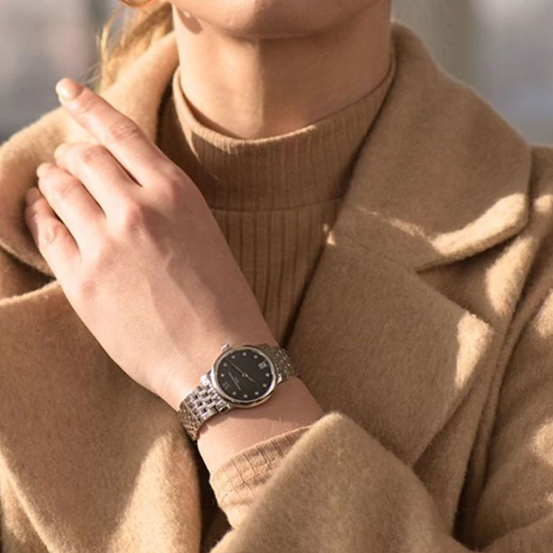 A person in a beige turtleneck and coat touches their neck, showcasing the Frederique Constant Slimline Quartz 30mm ladies' watch with a stainless steel bracelet and black face on their wrist.