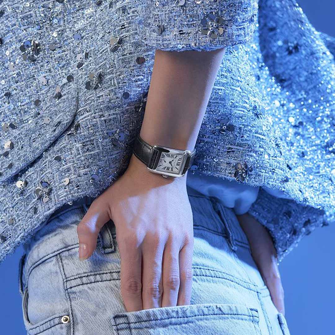 A person wearing a sparkly blue jacket and light denim jeans poses with one hand in their pocket, featuring the luxurious Frederique Constant Classics Carrée Small Seconds Quartz 36mm x 25mm watch. The black strap and square silver face exude timeless elegance, complemented perfectly by the solid blue background.