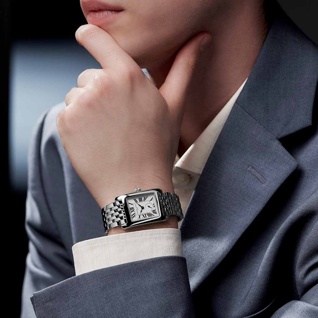 A person in a gray suit and white shirt rests their chin on one hand, highlighting the Frederique Constant Classics Carree Small Seconds Quartz 36mm x 25mm Watch with its silver metal band. The silvered dial adorned with Roman numerals captures an Art Déco elegance. The blurred background directs attention to this exquisite timepiece from Frederique Constant.