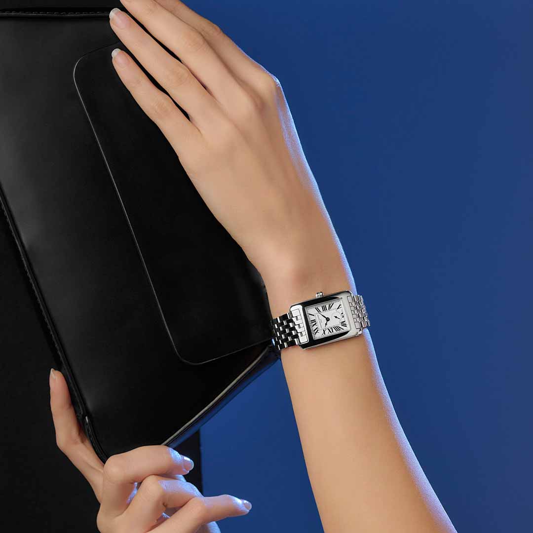 A person's hand wearing a Frederique Constant Classics Carree Small Seconds Quartz watch grasps a black tablet against a blue backdrop. The watch, featuring its 36mm x 25mm rectangular face and Roman numerals, embodies 1920s inspiration and Art Déco elegance.