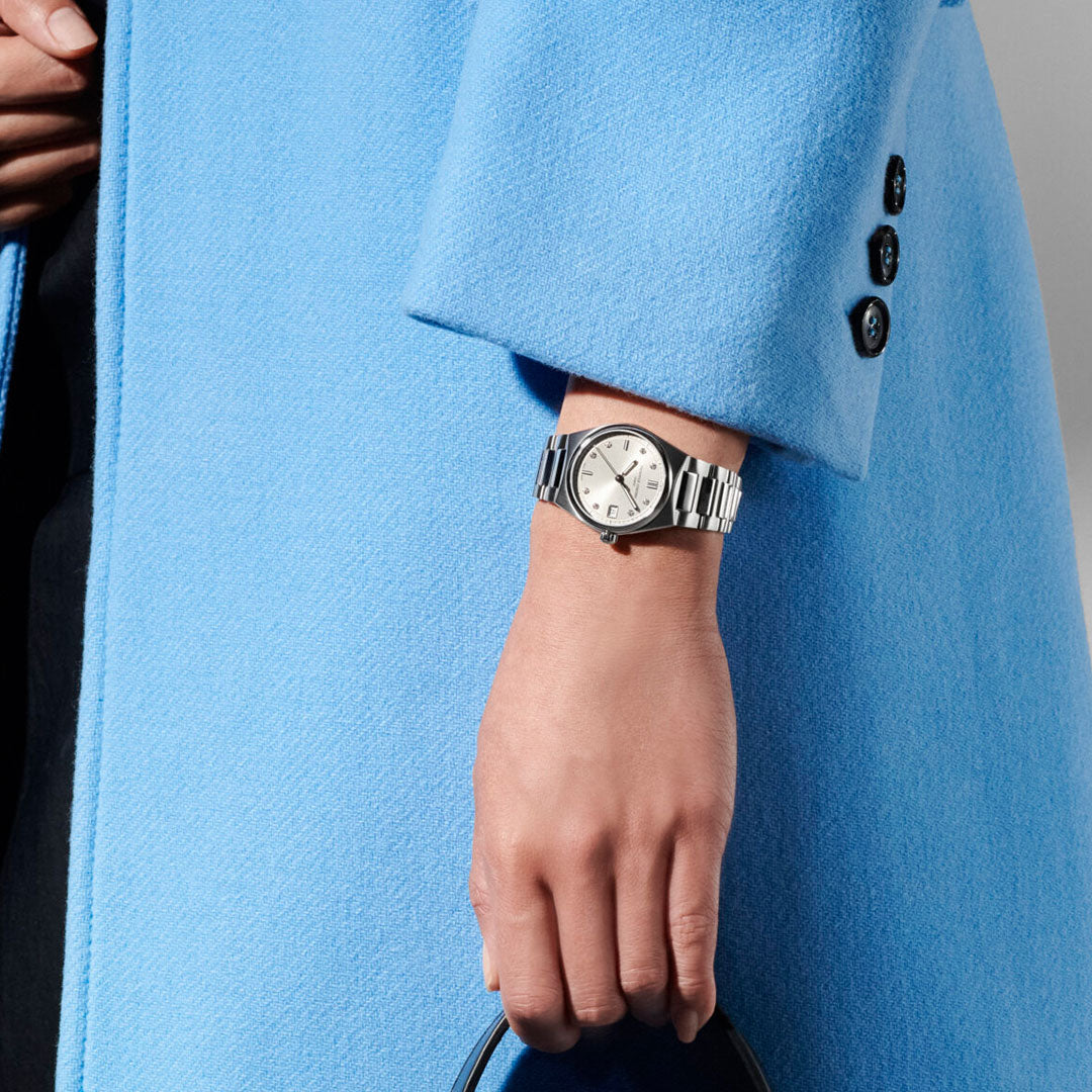 A hand wearing the Frederique Constant Highlife Ladies Quartz 31mm Watch with an interchangeable strap holds a black handle, shown with part of a light blue coat sleeve and black buttons visible.