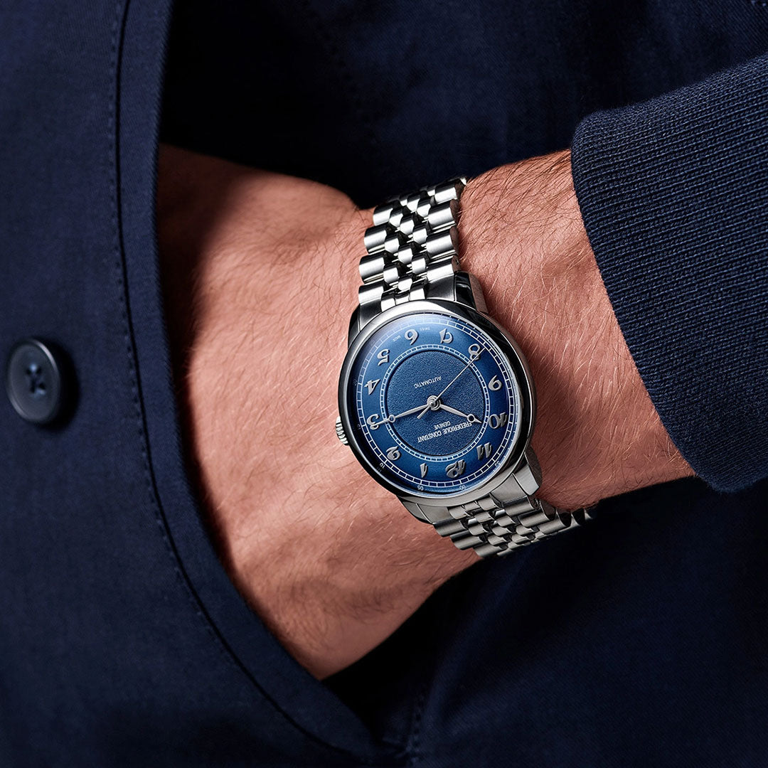A person in a dark blue shirt has their hand in their pocket, showing the Frederique Constant Classics Premiere Automatic 38.5mm Watch with a blue face, white numerals, a metal link band, and precise g100 la Joux-Perret movement.