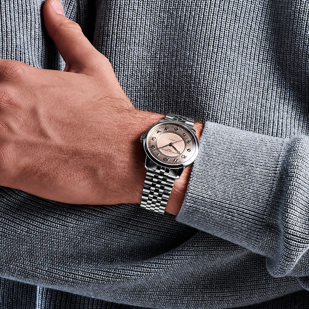A person wearing a Frederique Constant Classics Premiere Automatic 38.5mm watch with a pink dial and silver metal band, powered by the G100 La Joux-Perret movement, rests their hand on their body, dressed in a light gray ribbed sweater.