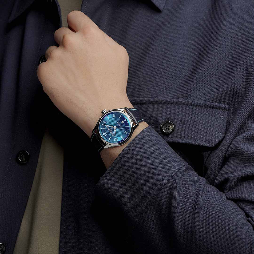 A person in a dark blue shirt crosses their arm over their chest, displaying a Geneva watchmaking masterpiece: the Frederique Constant Classics Premiere Automatic 38.5mm Watch. This exquisite timepiece features a striking blue face, complemented by a black leather strap, along with silver hour markers and hands, boasting an impressive 68-hour power reserve.