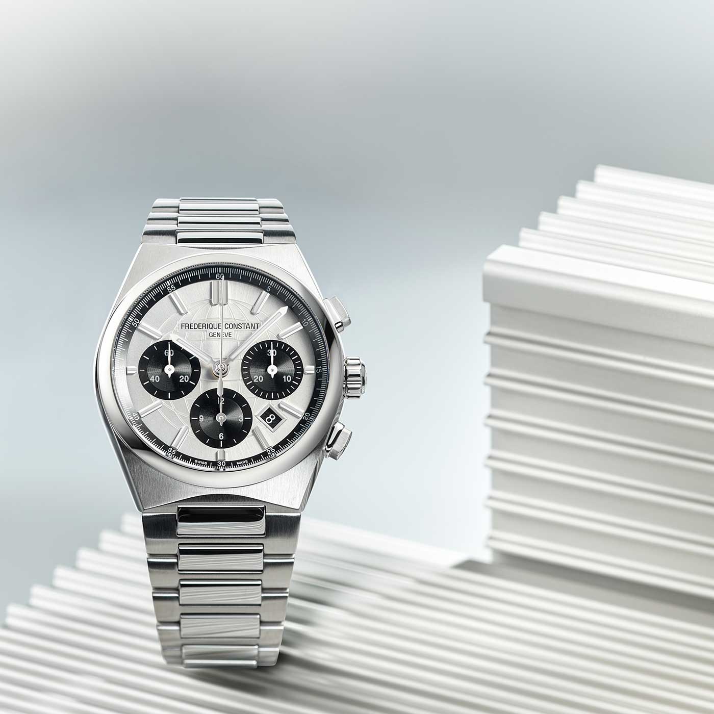 A Frederique Constant Highlife Chronograph Automatic 41mm watch with a stainless steel finish is displayed on ribbed white panels. The timepiece features a silver and black dial with three subdials, a date display, an anti-reflective sapphire crystal, and a coordinating metal bracelet. The background is soft and muted.