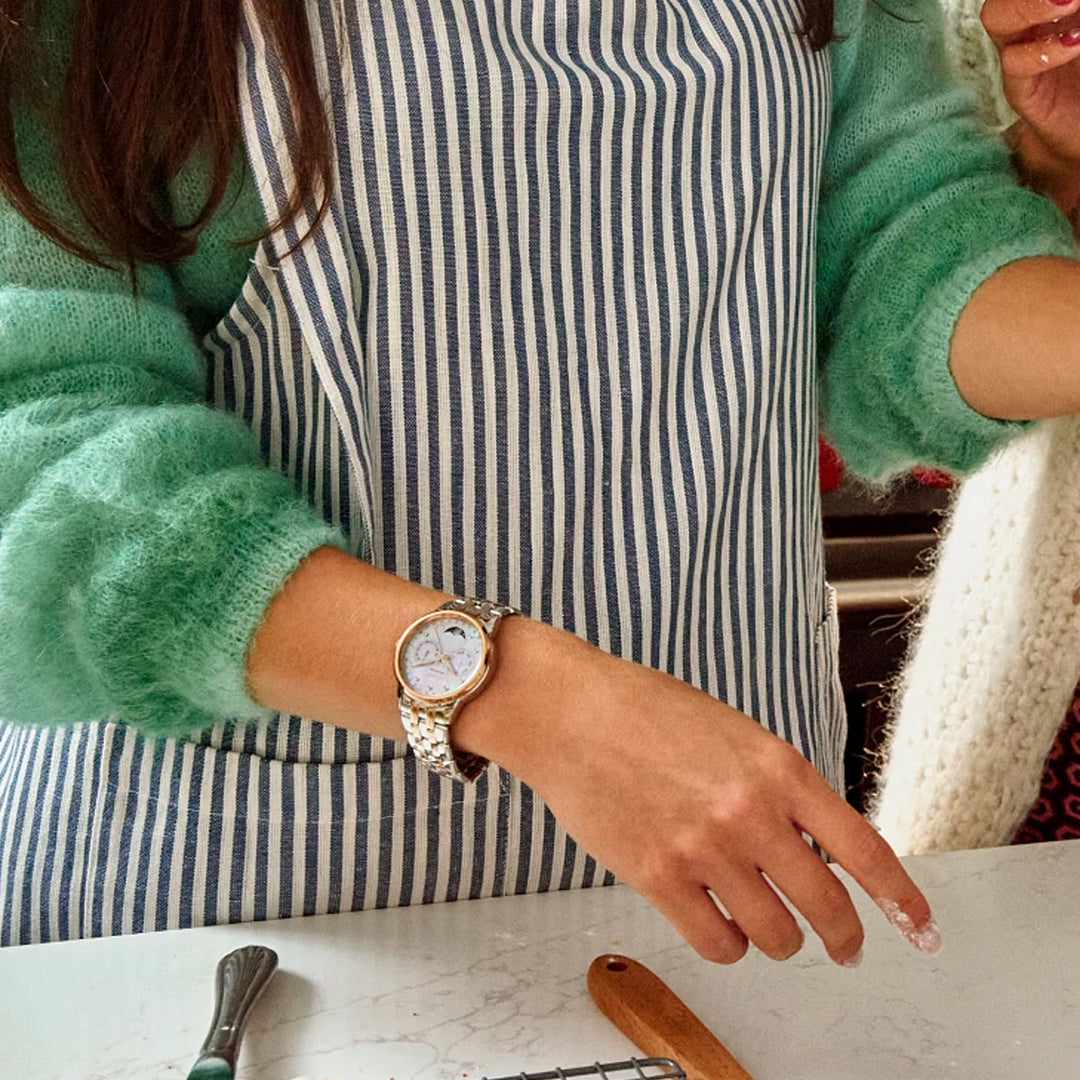 In the kitchen, someone in a green sweater and striped apron prepares food with various utensils around. Their wrist elegantly showcases a CITIZEN Calendrier Eco-Drive 37mm Watch, complete with a Mother-of-Pearl dial, bringing sophistication to the culinary scene.