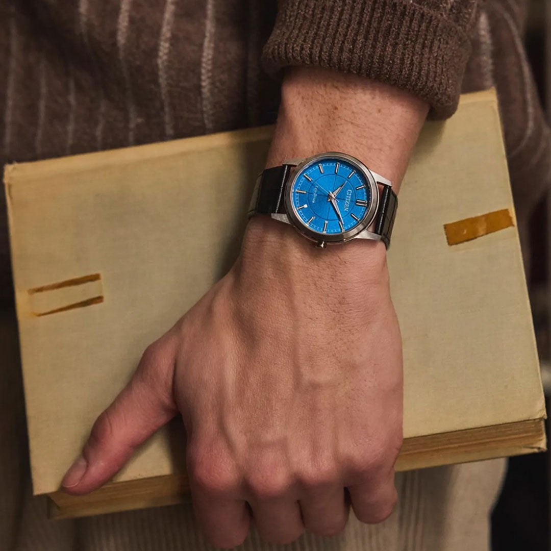 A person wearing the CITIZEN Rolan Eco-Drive 37mm Watch, featuring a stainless steel case and blue dial, holds a closed beige book marked by two old tape marks on the cover. The individual is dressed in a brown ribbed sweater.