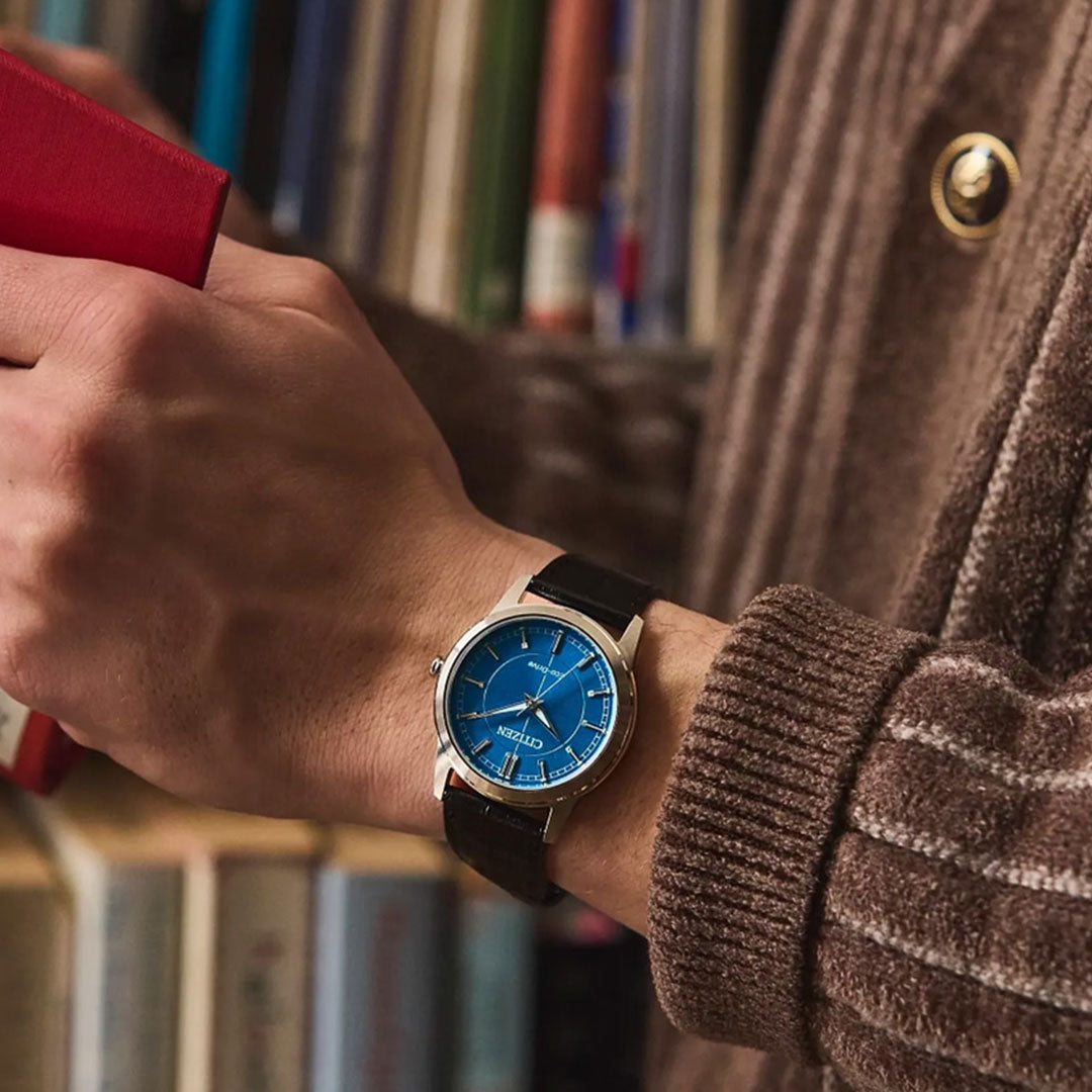 A person wearing a brown sweater and a CITIZEN Rolan Eco-Drive 37mm Watch holds a red book in front of a bookshelf filled with various books.