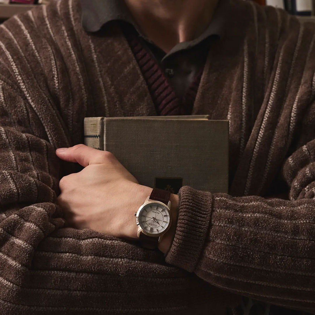A person in a brown, ribbed robe holds a closed gray book against their chest, arms crossed over it. On their wrist is the CITIZEN Rolan Eco-Drive 37mm Watch by CITIZEN, featuring an elegant mother-of-pearl dial. Their face remains out of view.