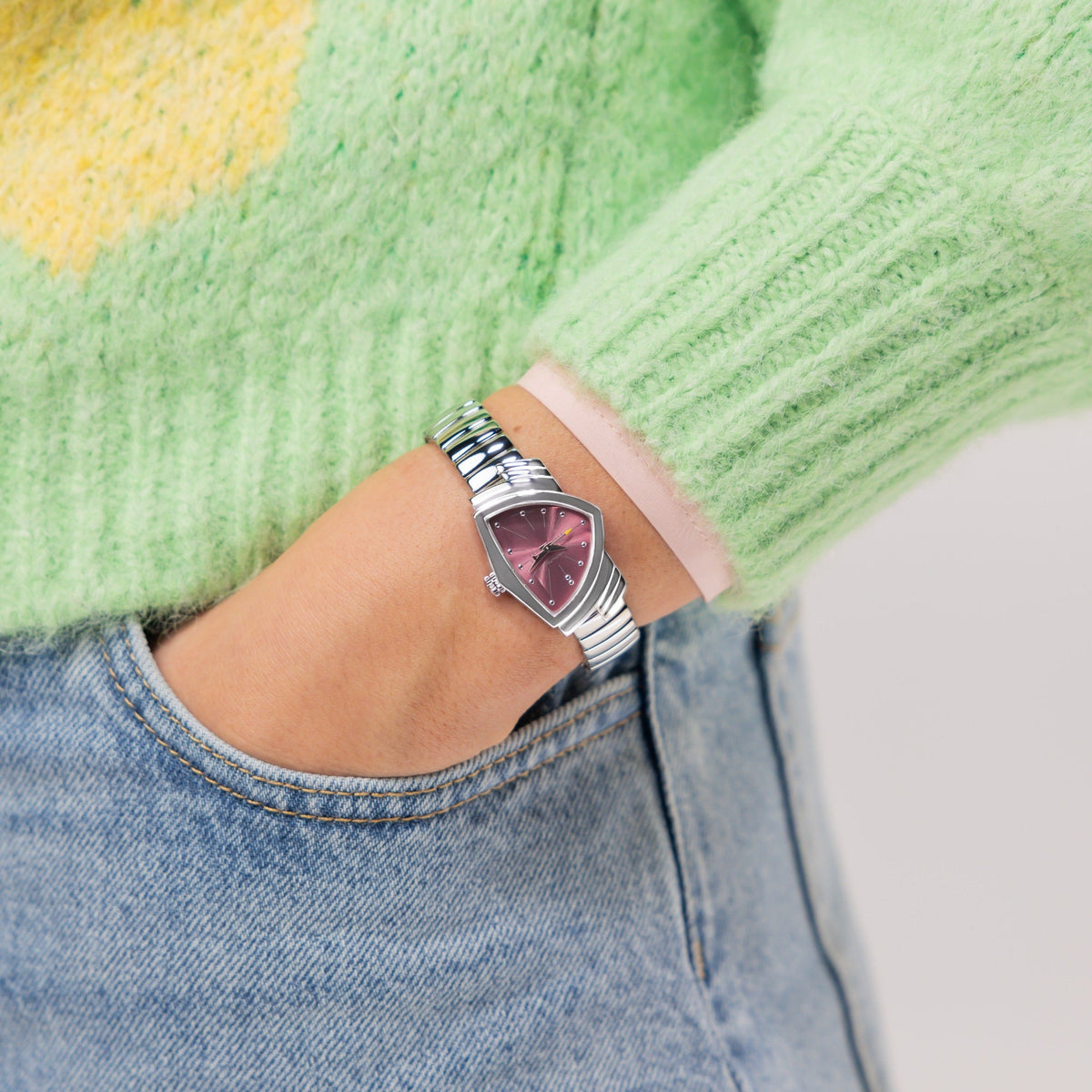 A person wearing a green sweater and blue jeans has their hand in their pocket, showcasing a Hamilton Watch Ventura S Quartz 24mm x 36mm timepiece featuring a purple, triangular dial and a sleek stainless steel bracelet.
