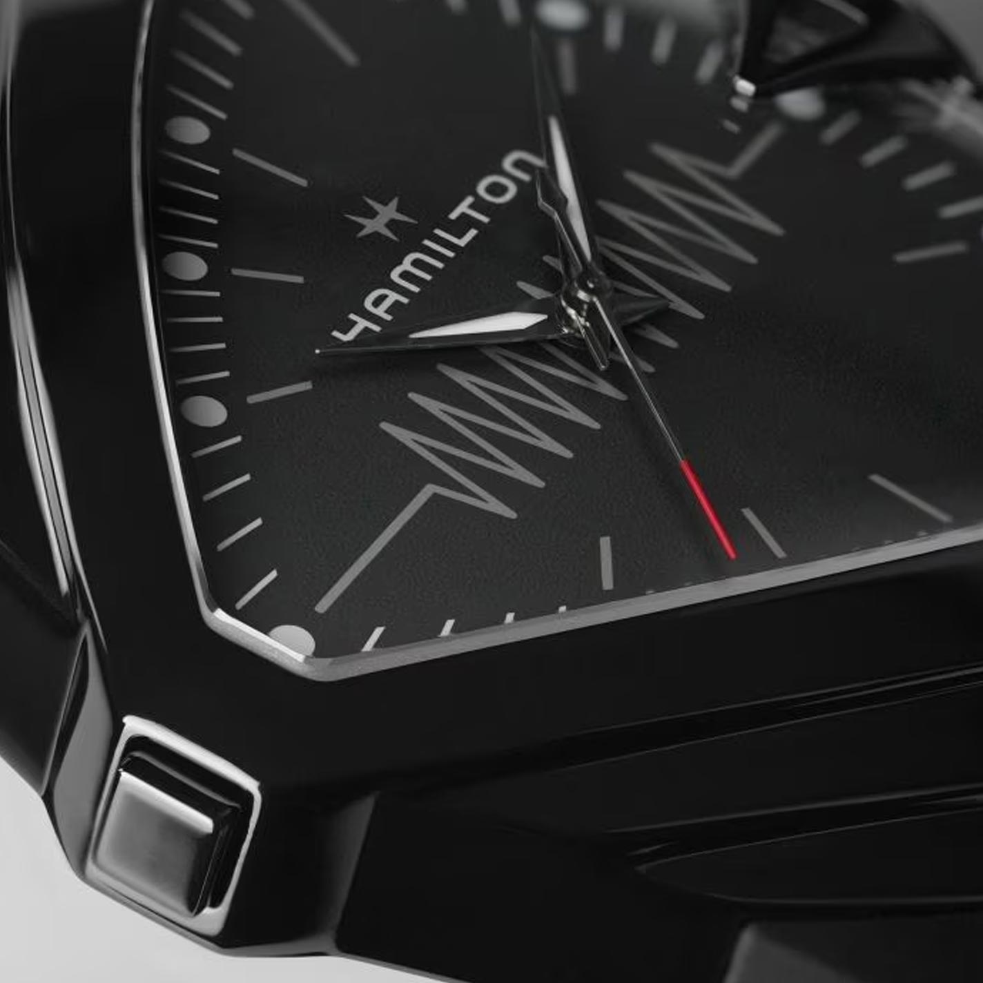 A detailed view of the Hamilton Ventura XXL Bright Quartz watch highlights a sleek black wristwatch with an angular design. The watch face proudly displays the Hamilton Watch logo, accompanied by silver hour markers and a subtle zigzag pattern. This battery-powered timepiece features silver hour and minute hands, with a distinctive red second hand for added contrast.