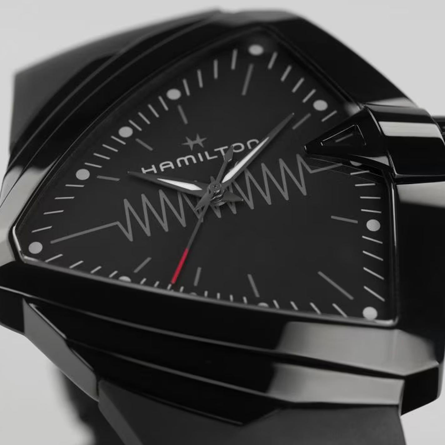 This close-up features the Hamilton Ventura XXL Bright Quartz 52mm X 47.6mm Watch, a stylish, battery-powered wristwatch with a bold triangular design. It showcases a zigzag pattern and a striking red-tipped second hand set against a glossy black casing, complemented by an eye-catching light-up dial.