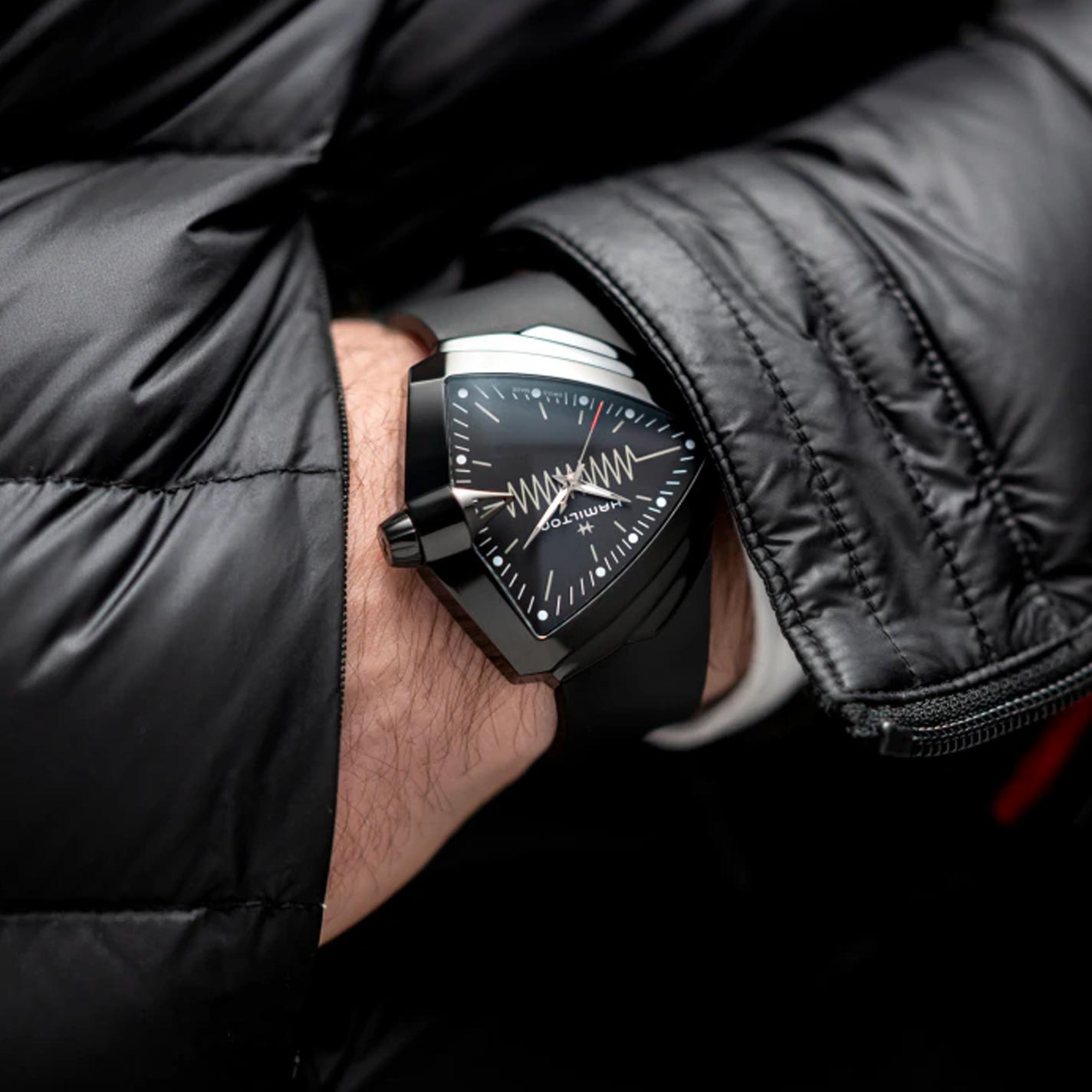 A person wearing a sleek Hamilton Ventura XXL Bright Quartz 52mm x 47.6mm watch with a zigzag pattern and a light-up dial from Hamilton Watch. They are also sporting a black quilted jacket, creating a stylish ensemble.