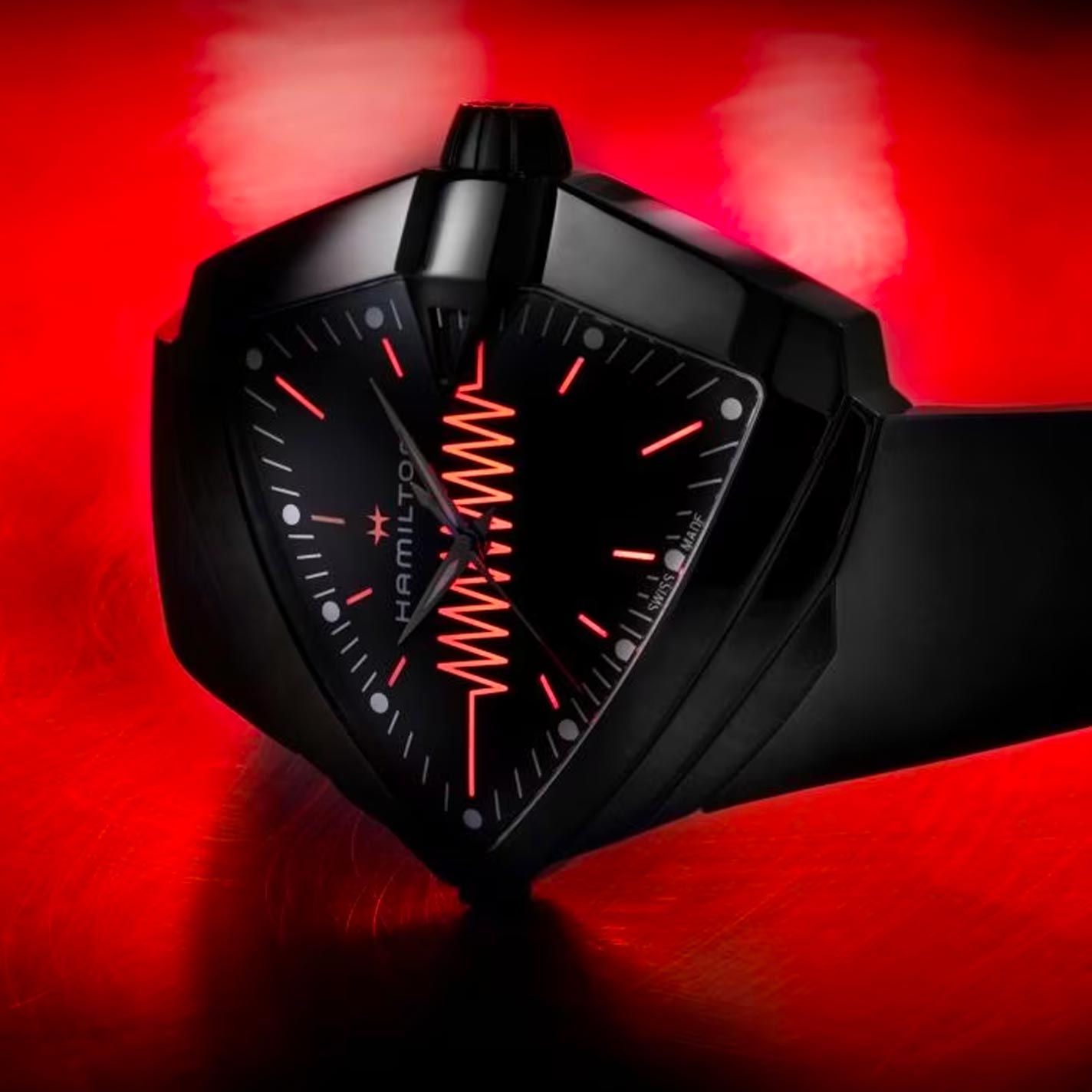 The Hamilton Ventura XXL Bright Quartz 52mm x 47.6mm Watch by Hamilton Watch is a futuristic timepiece with a black dial featuring a triangular face and red accents on a vibrant red backdrop. This battery-powered watch showcases unique markings and an angular black bracelet, offering a striking visual contrast.