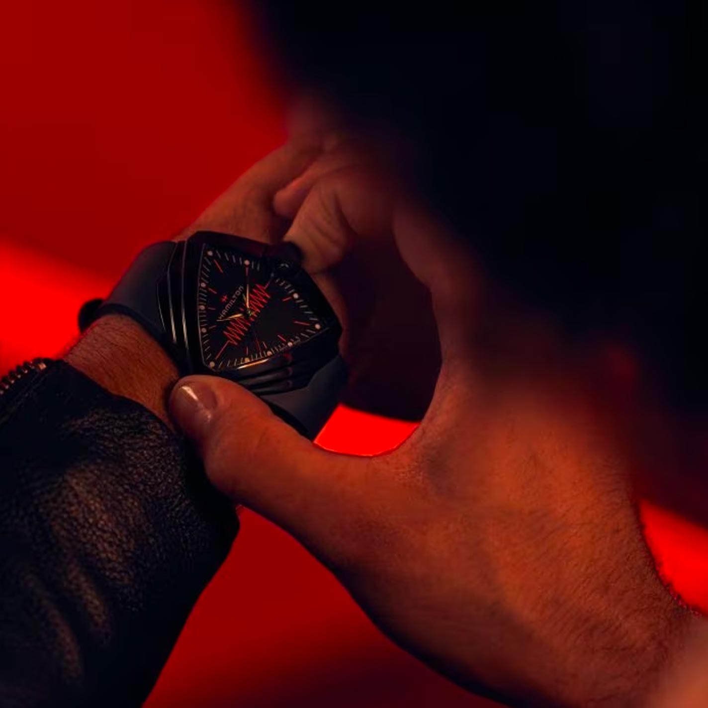 Clad in a leather jacket, an individual adjusts a Hamilton Ventura XXL Bright Quartz 52mm X 47.6mm Watch on their wrist against a vivid red background. This battery-powered timepiece from Hamilton Watch showcases a unique geometric design with striking red accents.