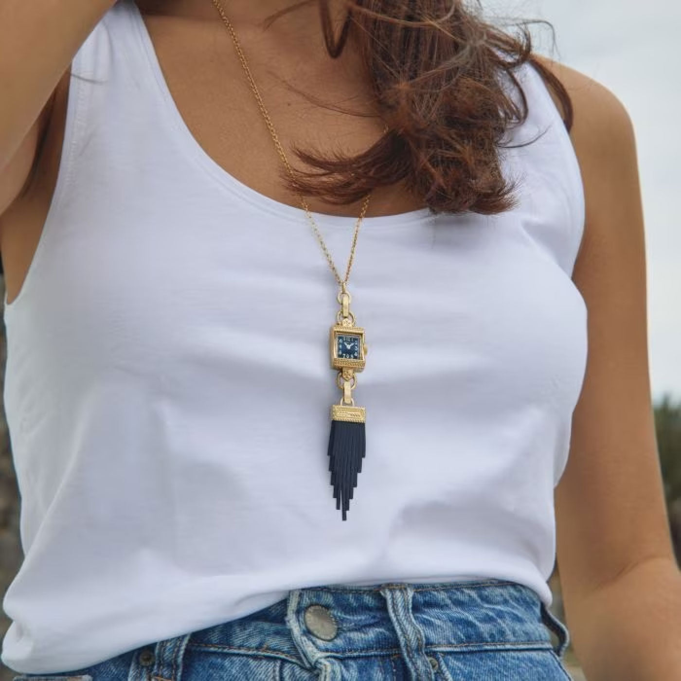 A person in a white sleeveless top and blue jeans is wearing a Hamilton American Classic Lady Hamilton Quartz 15mm x 19mm Watch Necklace by Hamilton Watch, featuring a long gold chain with a distinct blue pendant. The pendant showcases a square detail on top with an intricate, tiered fringe cascading below.