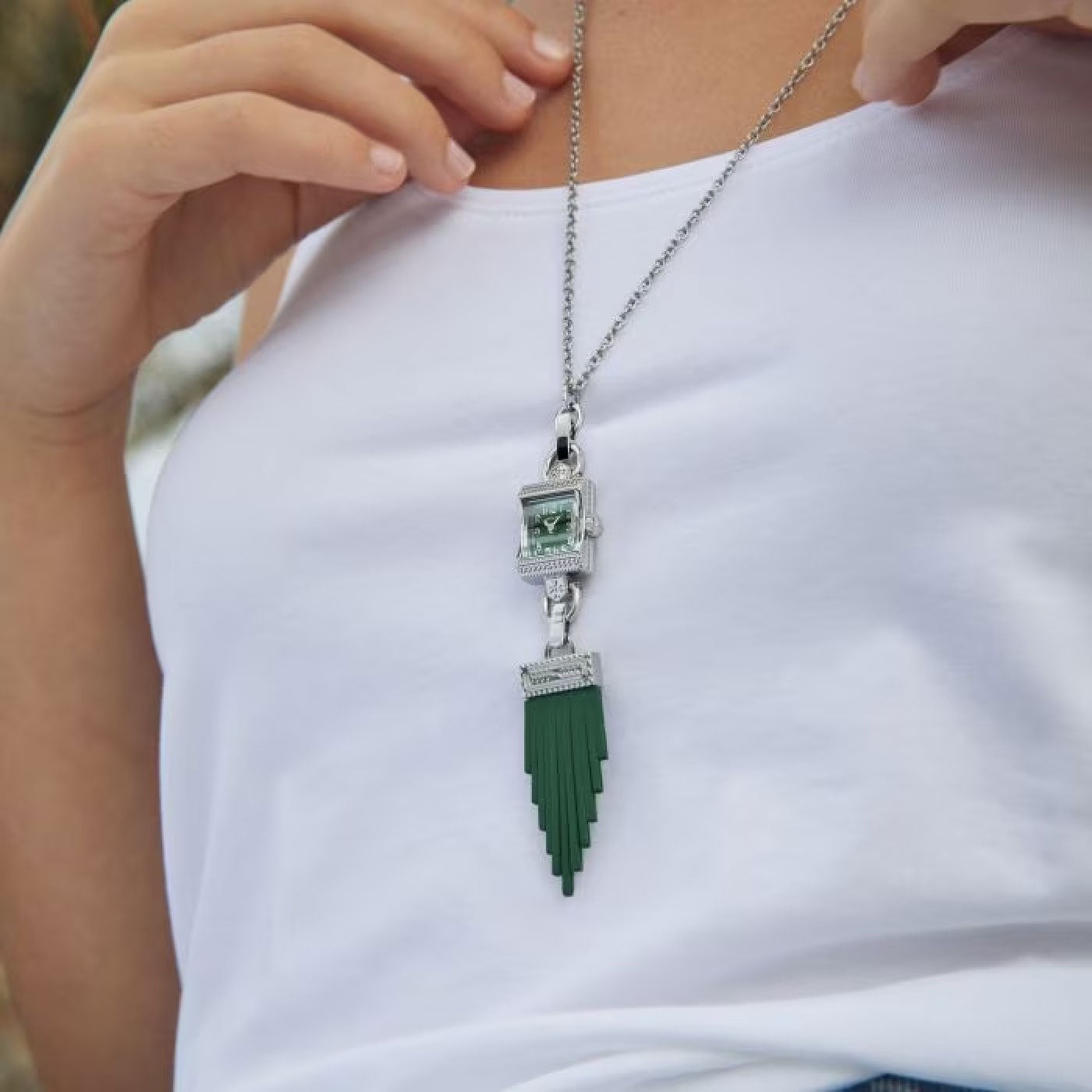 A person is wearing a white top accessorized with the Hamilton American Classic Lady Hamilton Quartz 15mm x 19mm Watch Necklace by Hamilton Watch, featuring a unique pendant with a green fringed ornament and garnet accents.