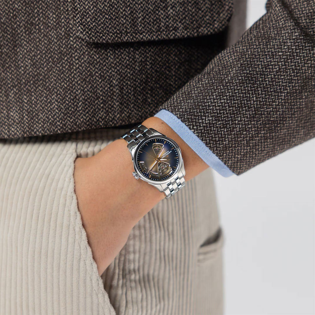 A person in a brown textured blazer and beige corduroy pants has their hand in their pocket, highlighting a Hamilton Jazzmaster Open Heart Lady Auto 36mm Watch by Hamilton Watch with a dark face on their wrist.