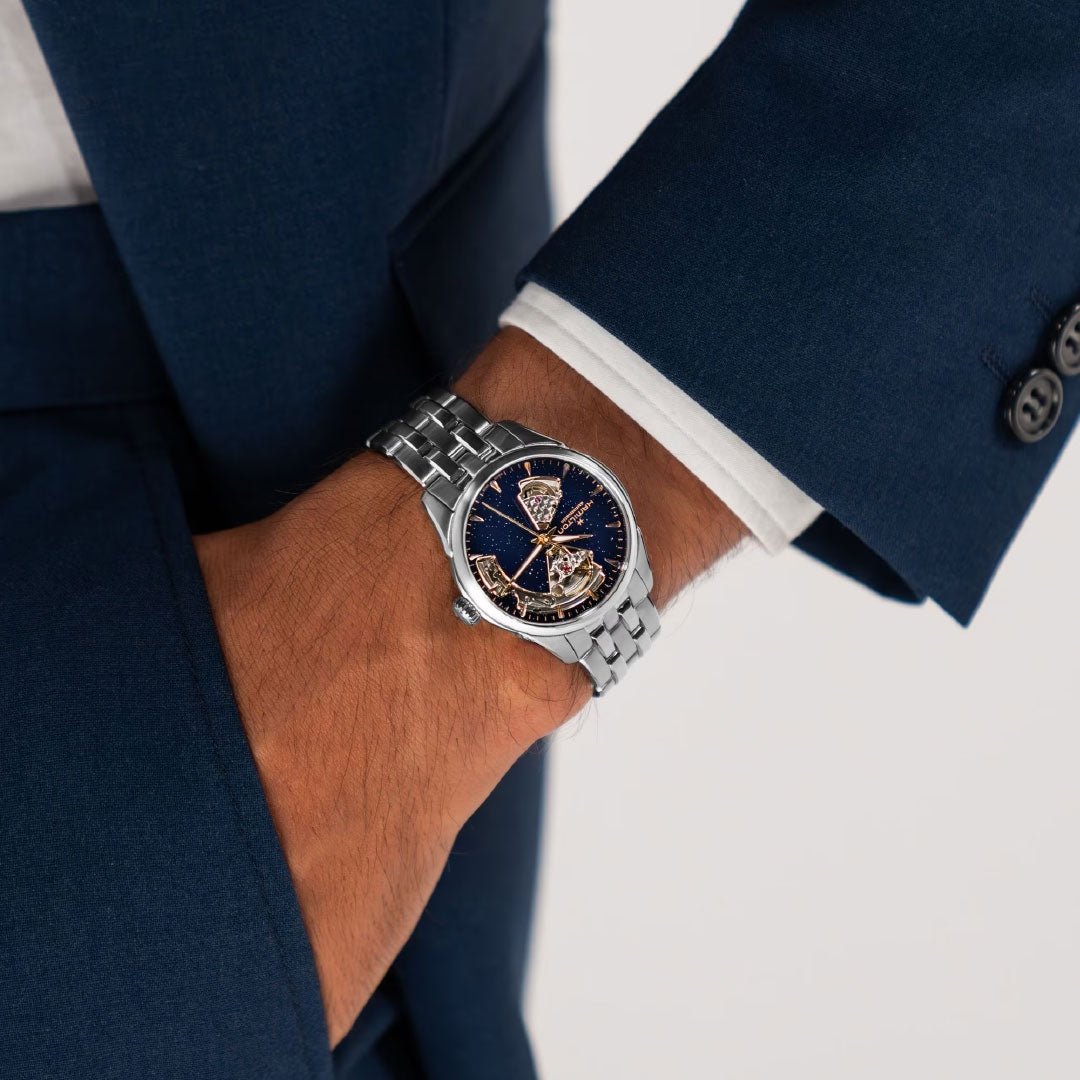 A person in a dark blue suit has their hand in their pocket, showing off the Hamilton Jazzmaster Open Heart Lady Auto 36mm Watch, which features a silver automatic movement and a detailed blue dial with gold accents.
