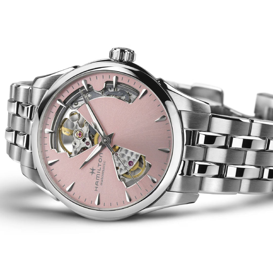 The Hamilton Jazzmaster Open Heart Auto 36mm by Hamilton Watch features a stainless steel band and a pink openwork dial showing the movement inside. Displayed at an angle on a white surface.
