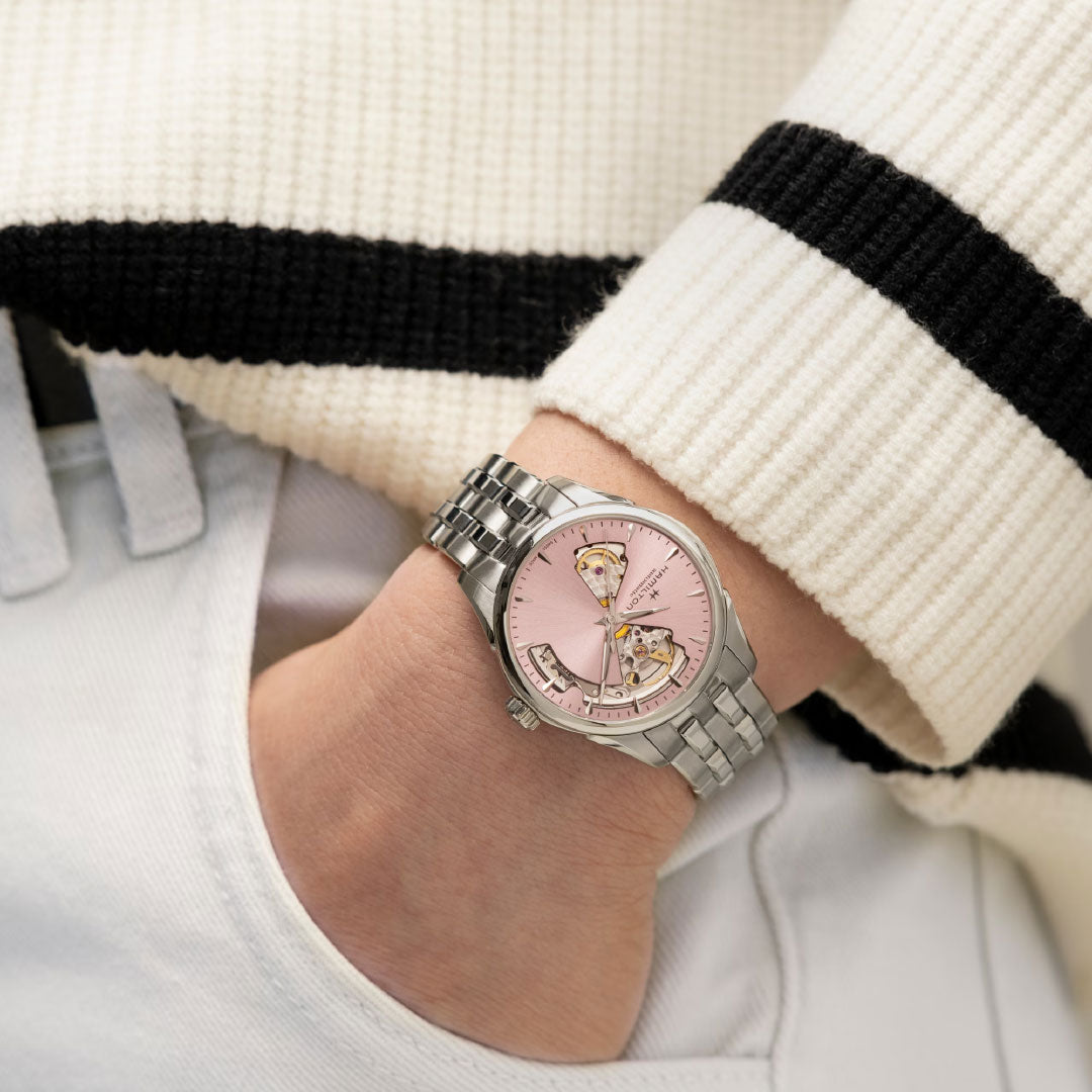 A person in a cream sweater with black stripes has their left hand in a pocket, displaying the Hamilton Jazzmaster Open Heart Auto 36mm Watch by Hamilton Watch, featuring a pink dial and heart-shaped cutouts.