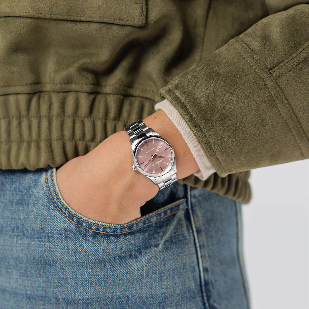 Wearing an olive green jacket and blue jeans, a person has their hand in their pocket while sporting a Hamilton Jazzmaster Quartz 32mm Watch with a pink face.