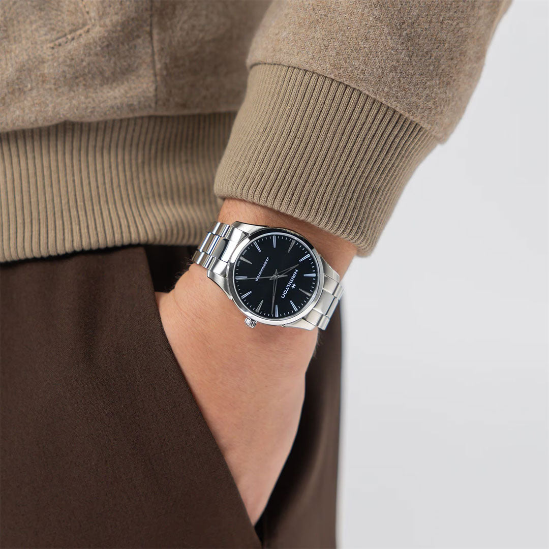 Wearing a beige sweater and brown pants, a person has their hand in their pocket, showcasing the Hamilton Jazzmaster Quartz 40mm Watch by Hamilton Watch with a sleek black dial and minimalist design.
