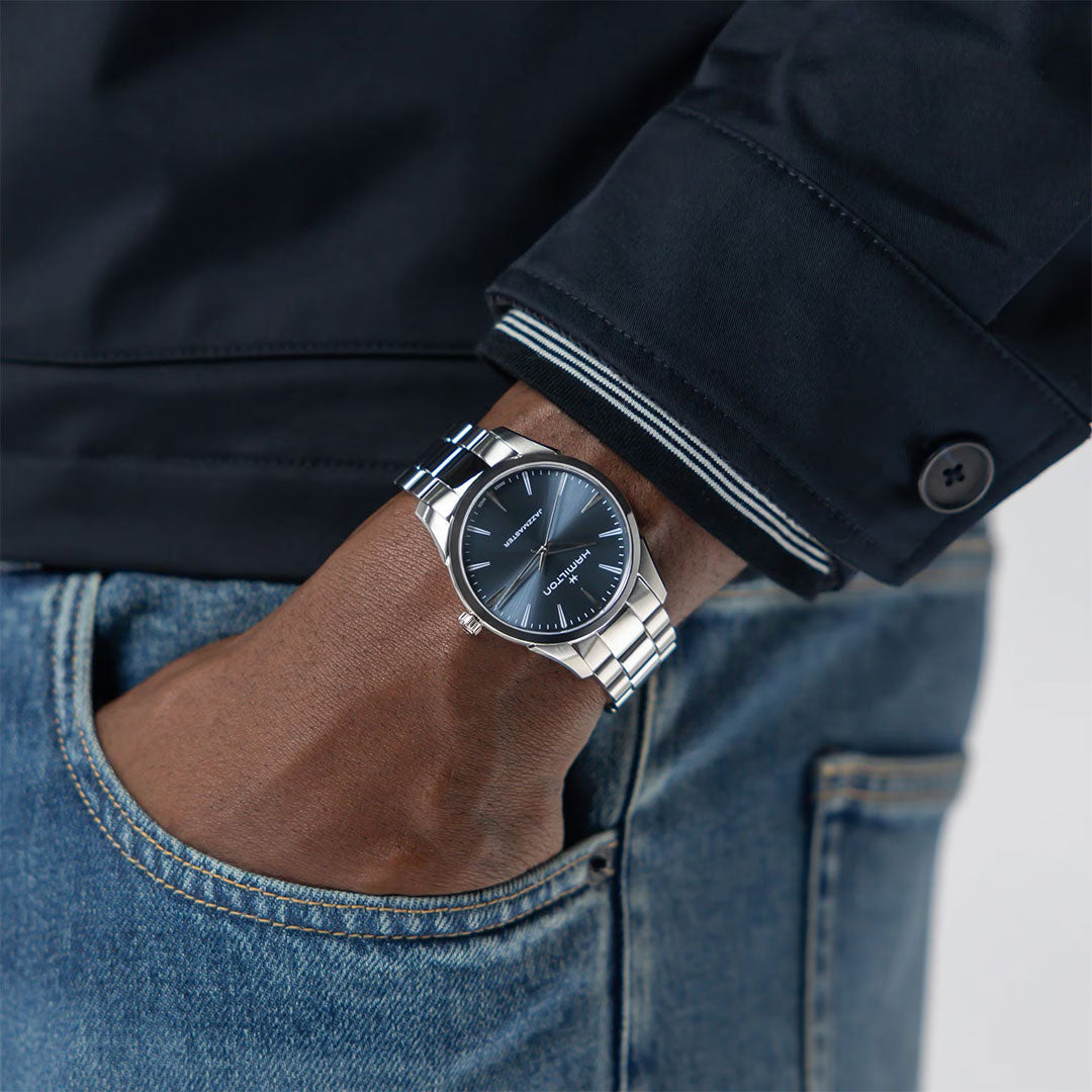 A person in a dark jacket and blue jeans has their hand in their pocket, displaying a stylish Hamilton Jazzmaster Quartz 40mm Watch with a blue dial and metal band on their wrist.