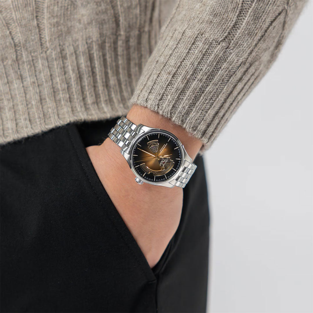A person in a gray knit sweater and black pants shows off the Hamilton Jazzmaster Open Heart Auto 40mm Watch by Hamilton Watch, featuring a gold case and brown leather strap.