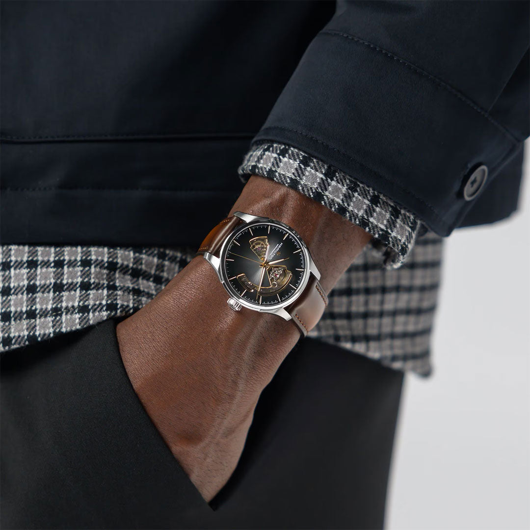 A person wearing a black and white checkered shirt, dark jacket, and black pants shows off a Hamilton Jazzmaster Open Heart Auto 40mm Watch by Hamilton Watch with a brown leather strap and black dial on their left wrist.