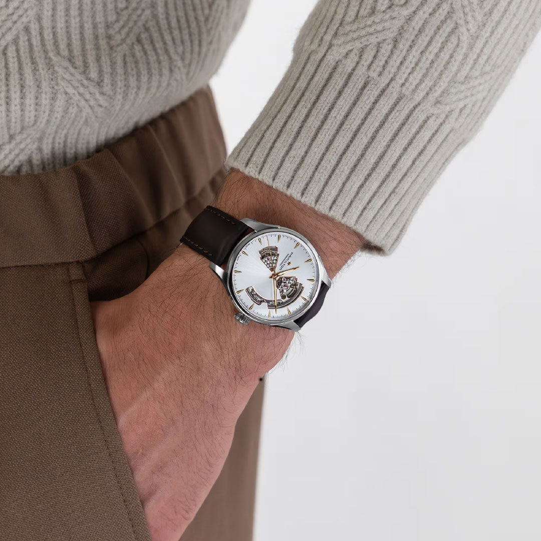 A person in a beige textured sweater and brown pants, hand in pocket, showcases the Hamilton Watch Jazzmaster Open Heart Auto 40mm with a brown leather strap and visible gears.