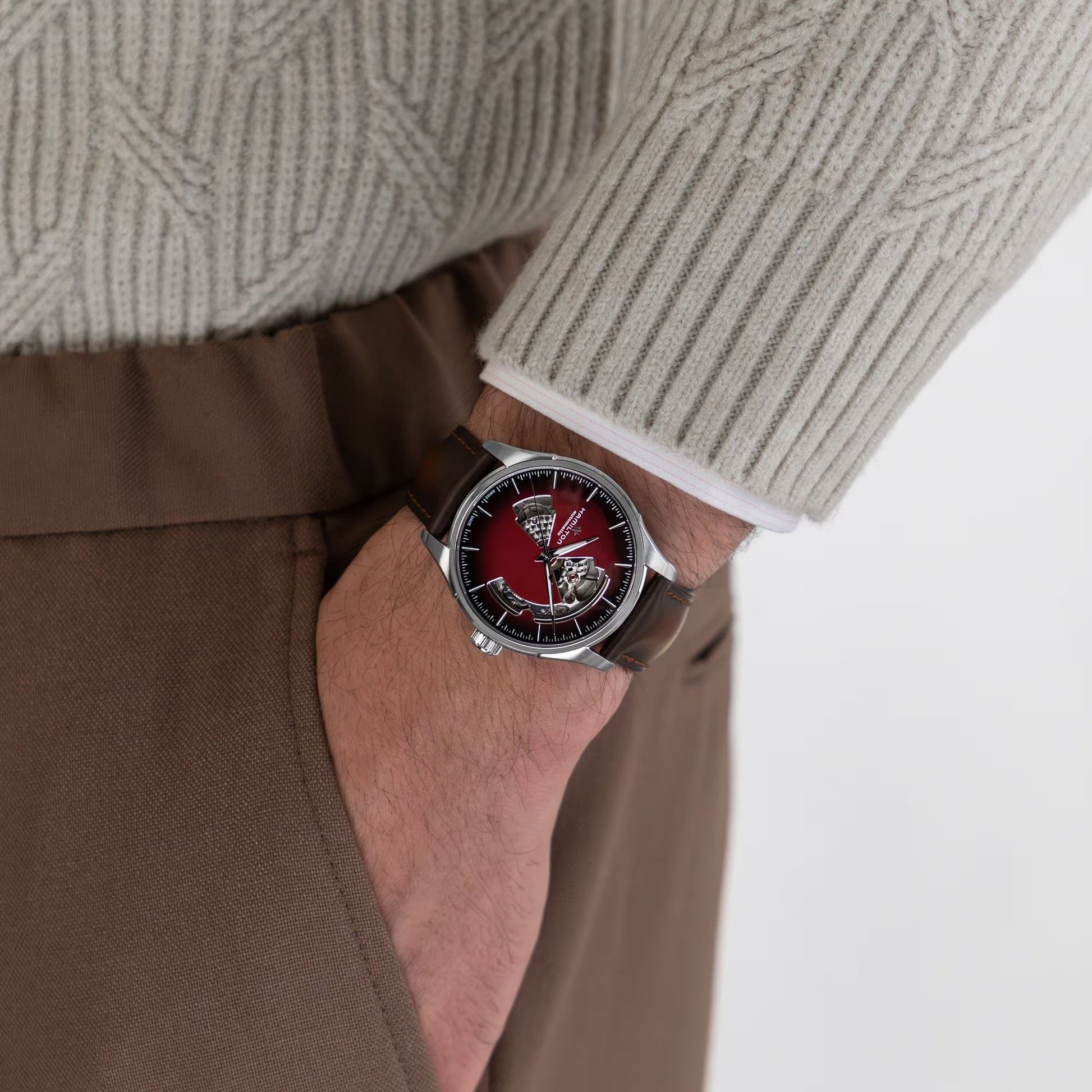 A person in brown pants and a beige knit sweater shows off a Hamilton Jazzmaster Open Heart Auto 40mm Watch by Hamilton Watch, featuring automatic movement and an elegant red dial on a brown leather strap.