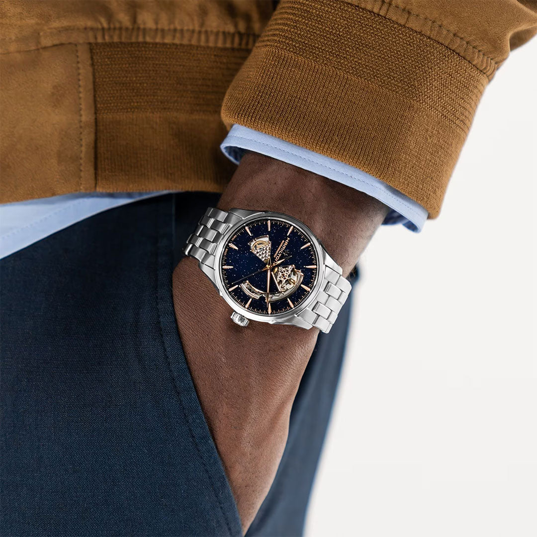 A person wearing a brown jacket and blue pants has their hand in their pocket, showing a Hamilton Jazzmaster Open Heart Auto 42mm Watch by Hamilton Watch with a blue and gold skeleton dial on their wrist.