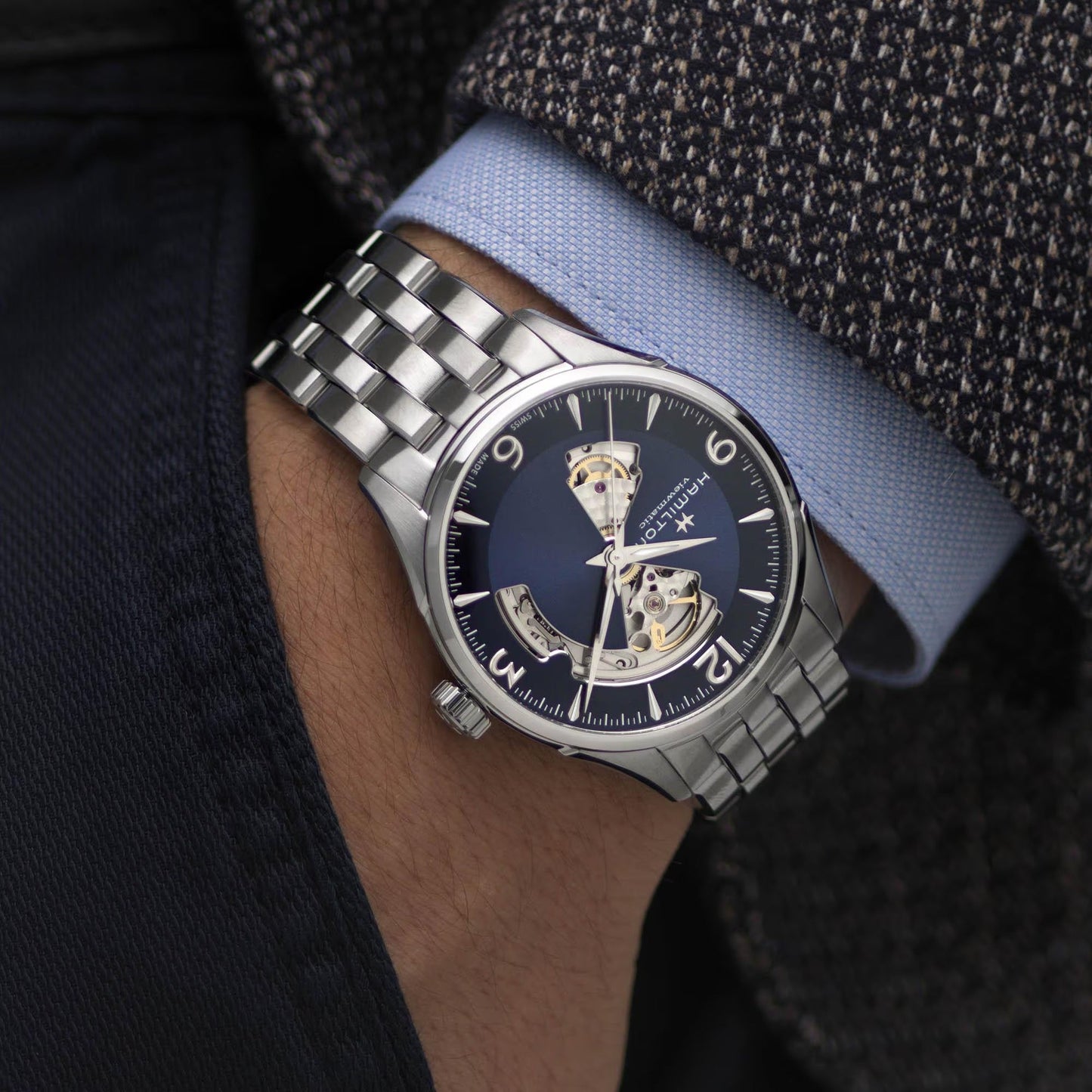 A person in a dark tweed blazer and light blue shirt, hand in pocket, wears the Hamilton Watch Jazzmaster Open Heart Auto 42mm with a blue dial and open-heart design.