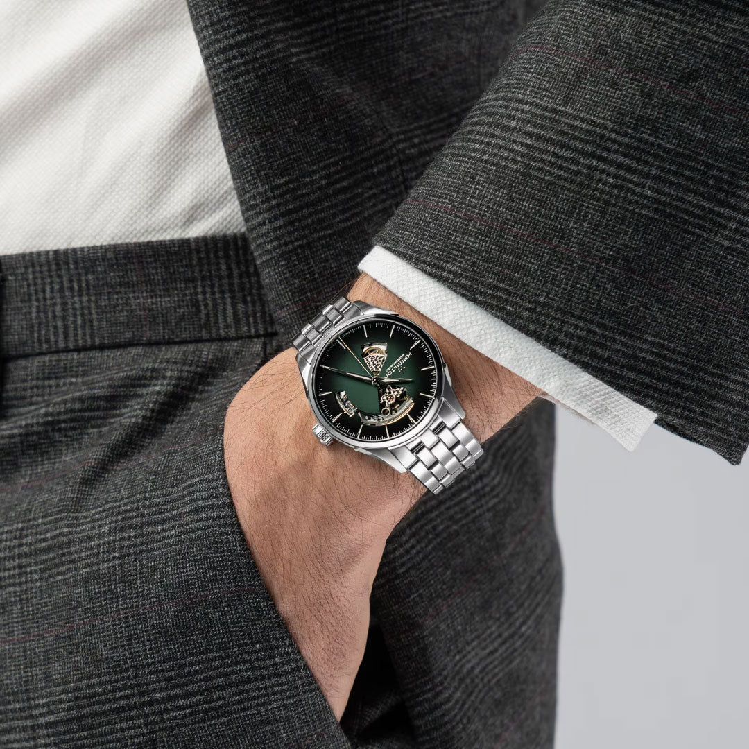 A person in a dark checkered suit with a white shirt has their hand in their pocket, showcasing the Hamilton Jazzmaster Open Heart Auto 42mm Watch by Hamilton Watch with a green face and visible mechanical movement.