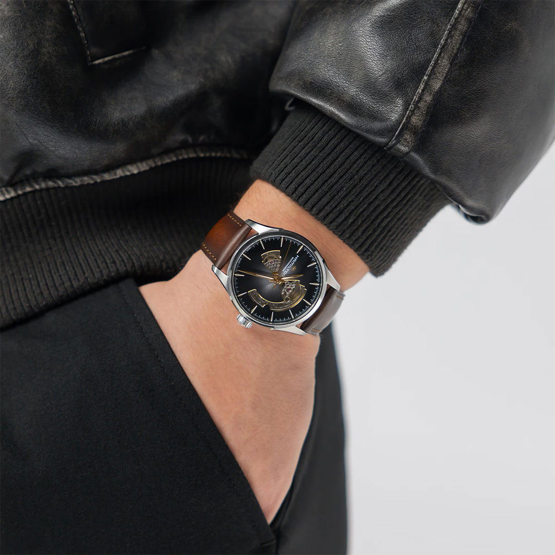 A person in a black leather jacket has their hand in their pocket, highlighting the Hamilton Jazzmaster Open Heart Auto 42mm Watch with a black dial, gold accents, and a brown leather strap by Hamilton Watch.