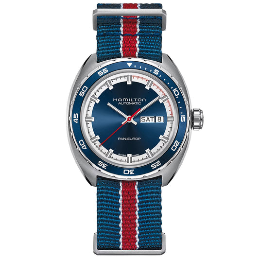 The Hamilton American Classic Pan Europ Day Date Auto 42mm Watch by Hamilton Watch exemplifies an American timepiece, boasting a blue face with a white dial, silver hands, and an automatic movement. The day-date display enhances its silver case and notched bezel, while the blue, white, and red striped NATO strap adds a distinct touch of style.