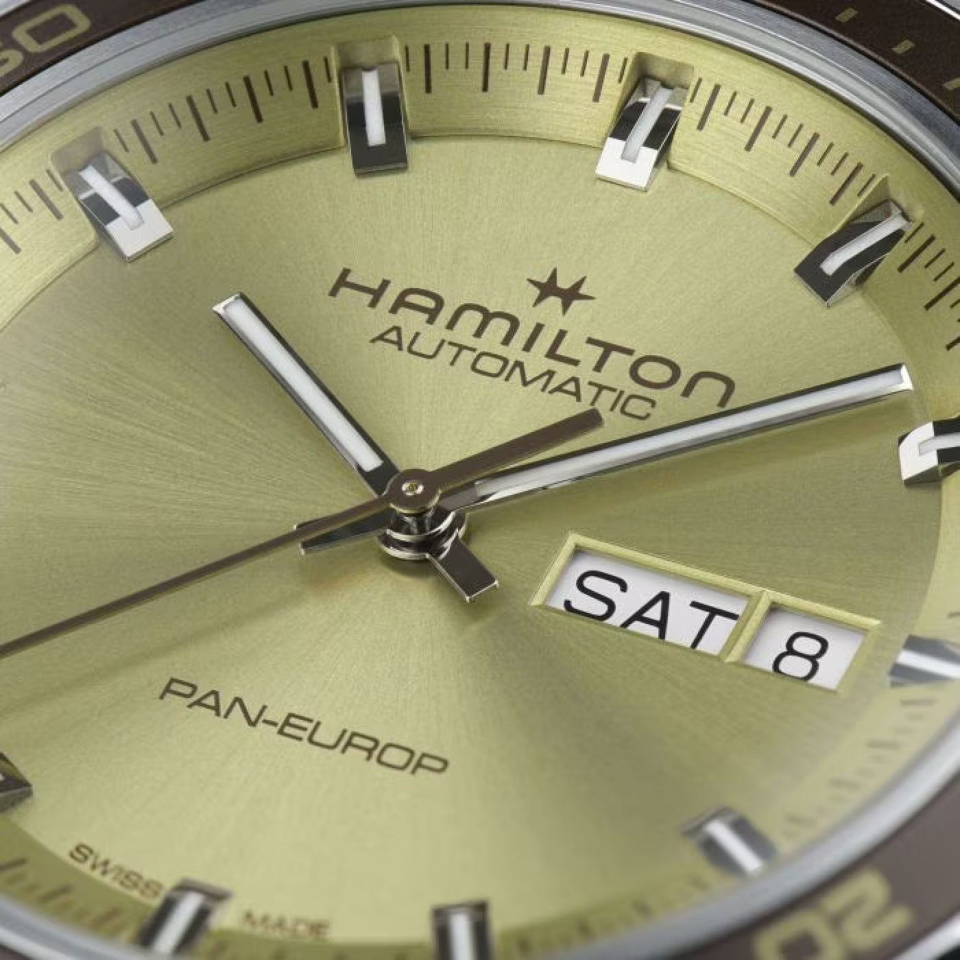 Close-up of a Hamilton American Classic Pan Europ Day Date Auto 42mm Watch featuring a captivating greenish-gold dial. The silver-toned hands and indices enhance the day-date display showing "SAT 8." The watch face prominently features the Hamilton Watch logo and "Swiss Made" text at the bottom, showcasing its exquisite craftsmanship.