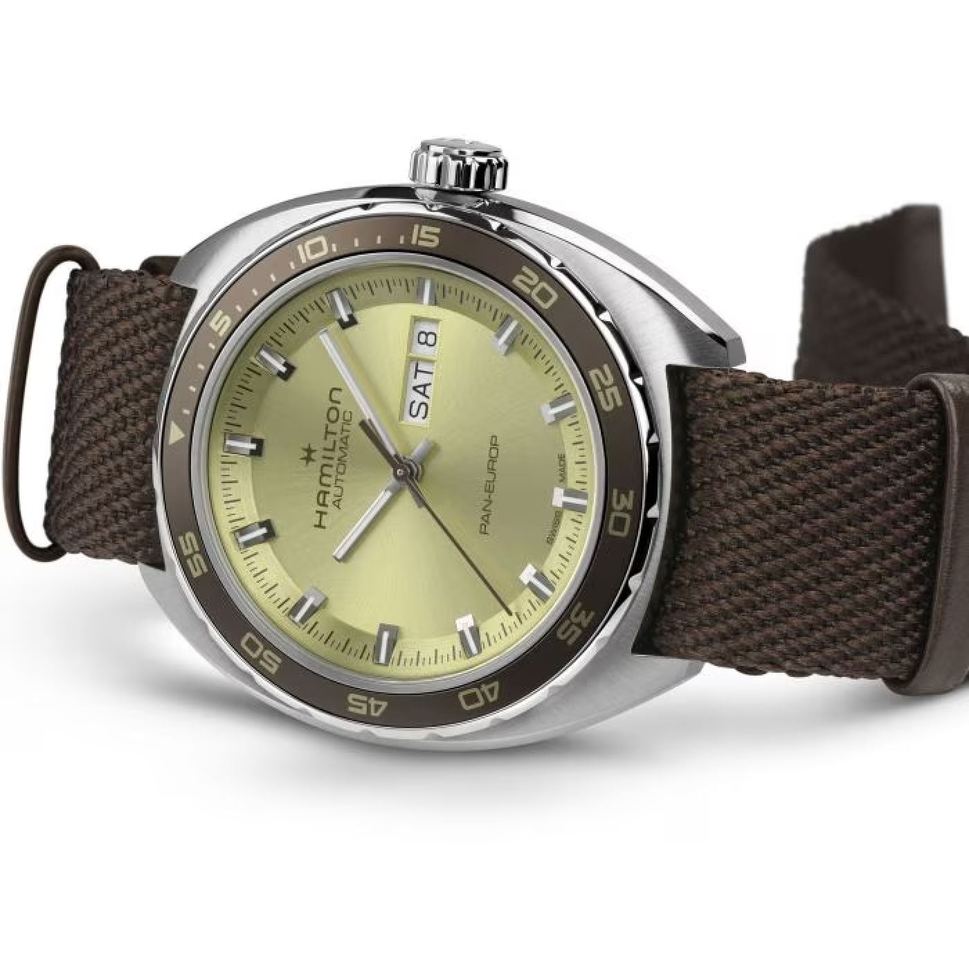 The Hamilton American Classic Pan Europ Day Date Auto 42mm Watch boasts a sophisticated green dial complemented by a brown fabric strap and a silver bezel with distinct hour markers and numbers. Additionally, the day and date are conveniently displayed at the 3 o'clock position, with the distinguished "Hamilton Watch" brand name prominently featured on the dial.