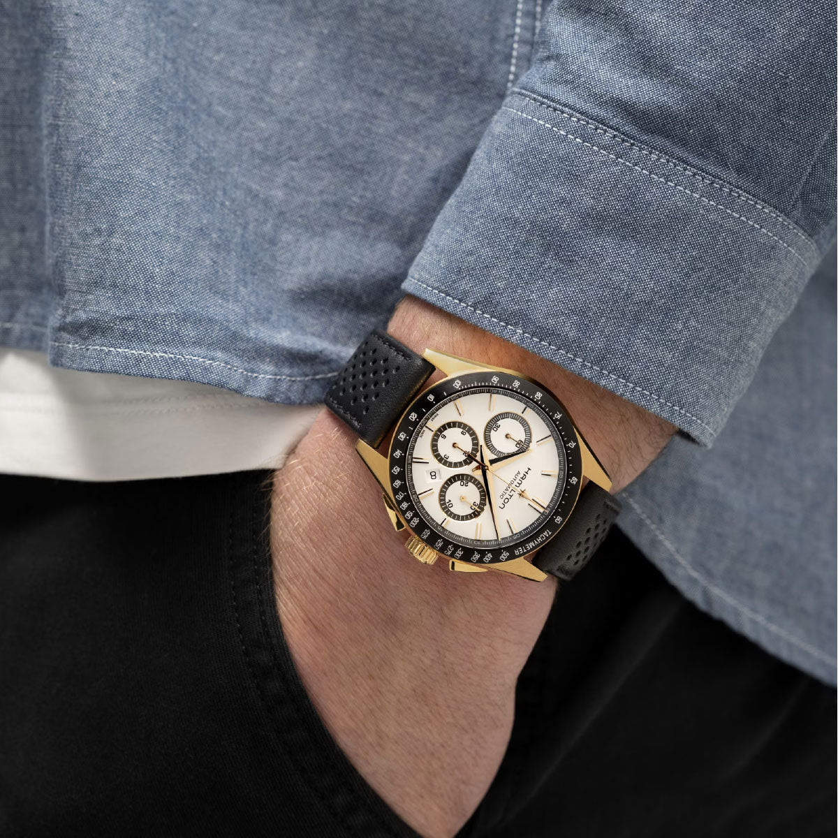 A person wearing the Hamilton Jazzmaster Performer Auto Chrono 42mm Watch by Hamilton Watch, a sophisticated gold and black automatic chronograph wristwatch with a white dial and multiple sub-dials. Paired with a light blue shirt and black pants, their look exudes sport-chic elegance as they casually slip one hand into their pocket.