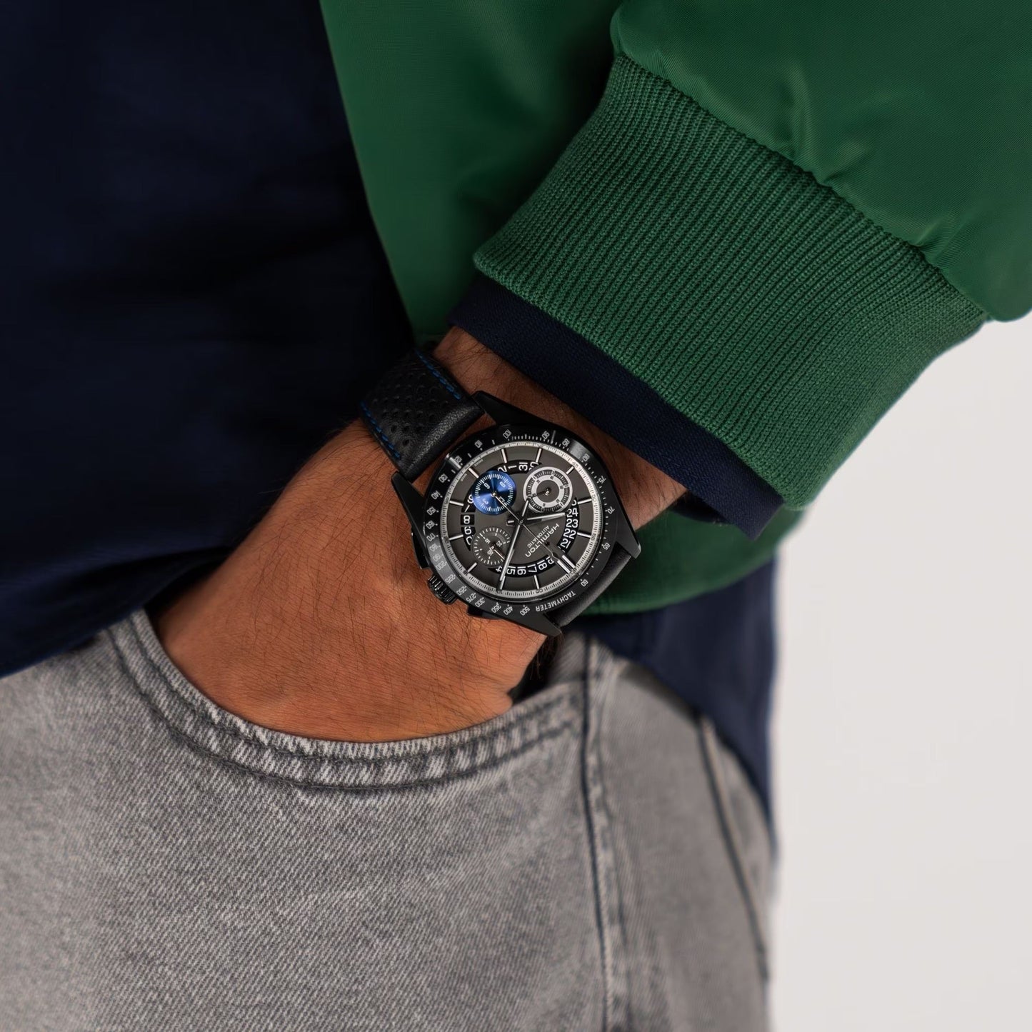 A person wearing a Hamilton Jazzmaster Performer Auto Chrono 42mm watch with a black leather strap, dressed in a green and navy jacket and light gray jeans, has their hand in their pocket.