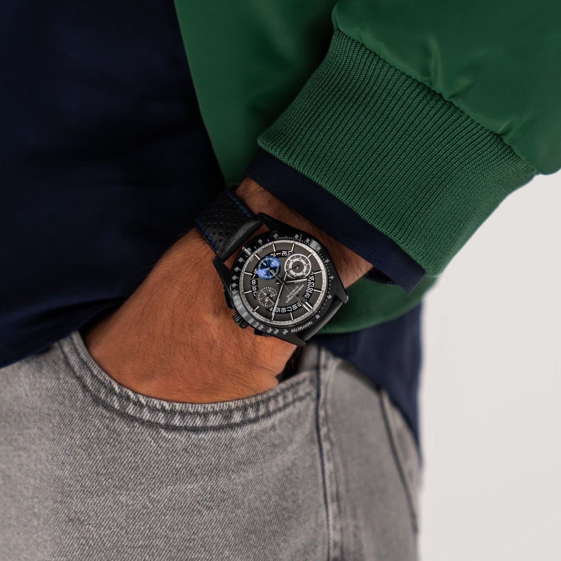 A person wearing a Hamilton Jazzmaster Performer Auto Chrono 42mm watch with a black leather strap, dressed in a green and navy jacket and light gray jeans, has their hand in their pocket.