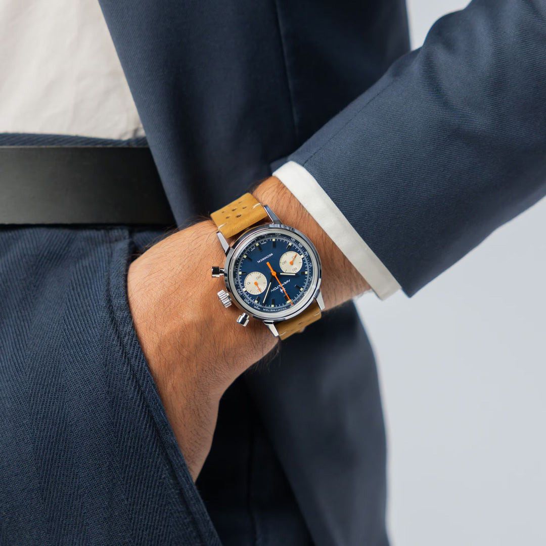A person in a dark blue suit with their hand in their pocket showcases the Hamilton American Classic Intra-Matic Mechanical Chronograph H 40mm Watch, featuring a blue dial, tan leather strap, three subdials, and striking red hands.