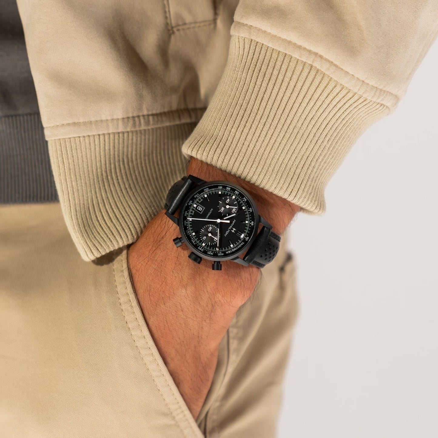A person in a beige jacket and pants, hand in pocket, sports the Hamilton American Classic Intra-matic Auto Chrono 40mm Watch by Hamilton Watch—vintage-inspired with racing roots, a black round face, and multiple dials.
