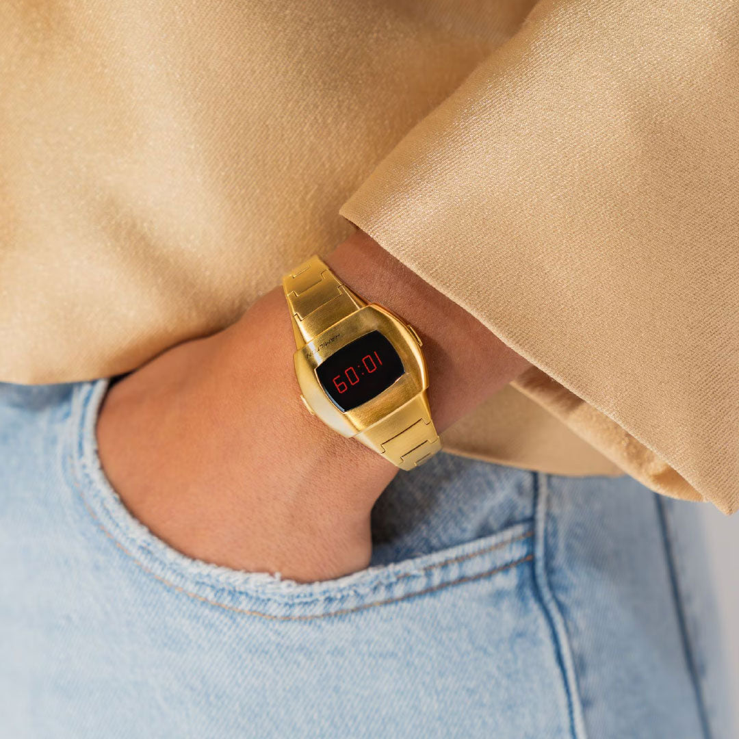 A person in light blue jeans and a beige top, hand in pocket, wears a Hamilton American Classic PSR 74 Quartz 26mm x 31mm Watch by Hamilton Watch, its red LED screen displaying "6:00".