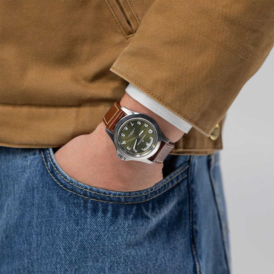 A person in blue jeans and a brown jacket, left hand in pocket, showcases the Hamilton Khaki Field King Auto 40mm Watch by Hamilton Watch with a brown leather strap and black dial.