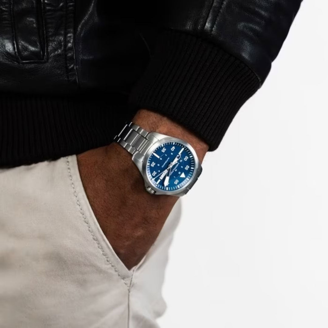 A person sporting a black leather jacket and beige pants has their left hand casually tucked into their pocket, proudly displaying a Hamilton Khaki Aviation Pilot Day Date Auto 42mm Watch. This elegant wristwatch from Hamilton Watch features an automatic movement, complemented by a striking blue dial, silver hour markers, and a sleek metal band. The scene is set against a plain white background.