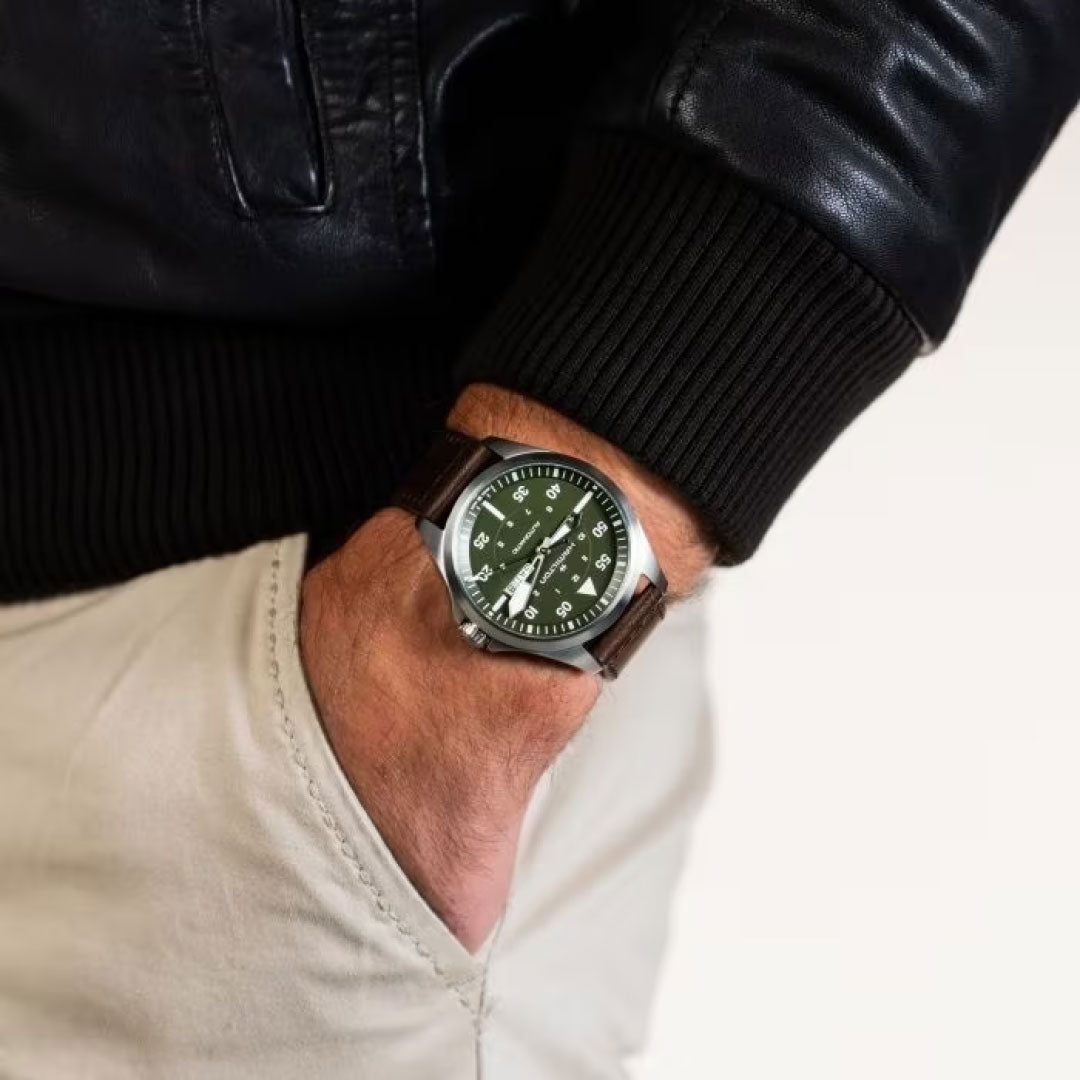 A person dressed in a black leather jacket and beige pants casually places their hand in their pocket, displaying the Hamilton Khaki Aviation Pilot Day Date Auto 42mm Watch. This watch, from the Hamilton Watch brand, features a brown leather strap and a green dial with automatic movement, elegantly highlighting casual yet stylish fashion.