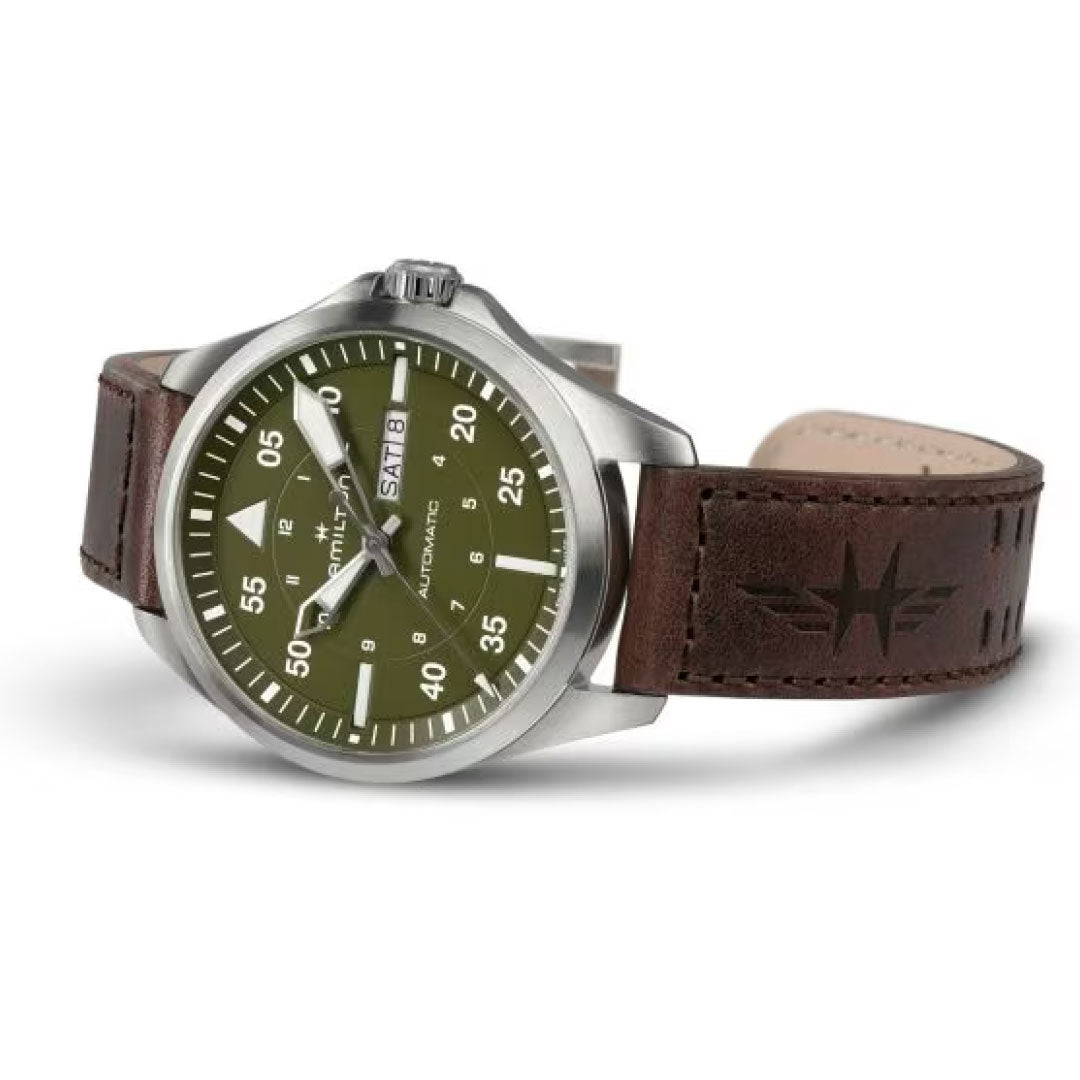 The Hamilton Khaki Aviation Pilot Day Date Auto 42mm Watch by Hamilton Watch features a brown leather strap with an aviator logo. It boasts a green dial with white numerals and a silver case, showcasing a rugged, classic design complemented by automatic movement for precision.