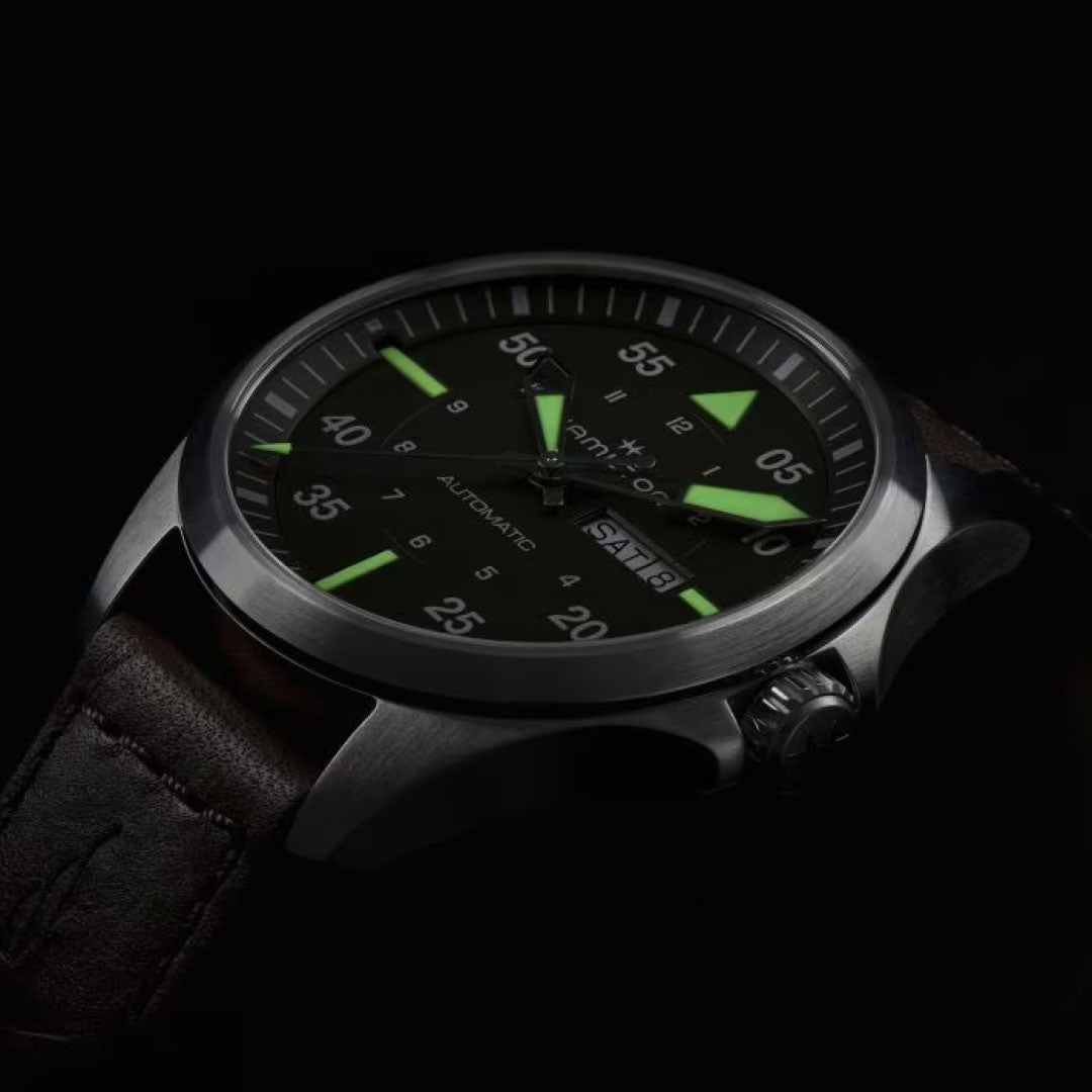 The Hamilton Watch brand presents the Khaki Aviation Pilot Day Date Auto 42mm watch, showcasing its dark dial with luminescent green hands and markers. The metallic case and leather strap emphasize the automatic movement of this timepiece, all set against a sleek black backdrop.